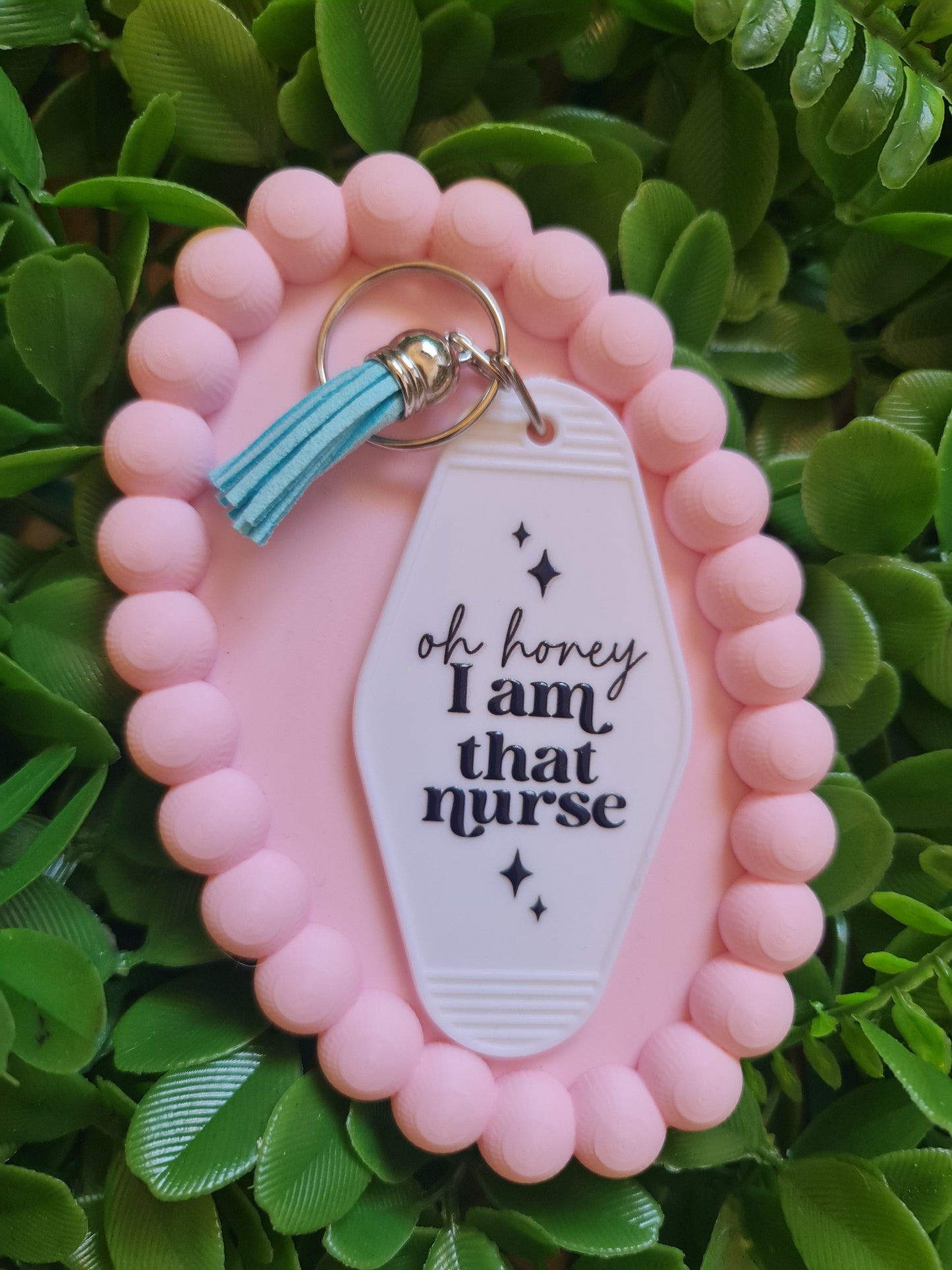 Pink keychain with a white tag and turquoise tassel on a green leafy background