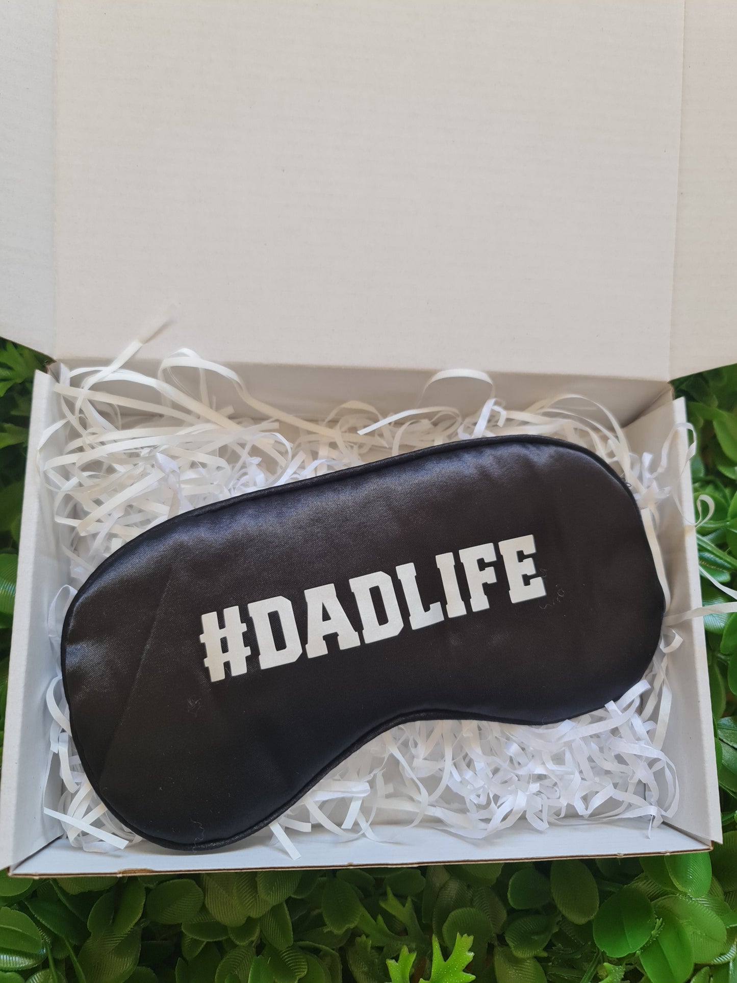 Black sleep mask with '#DADLIFE' text in a box with white tissue paper, surrounded by greenery.