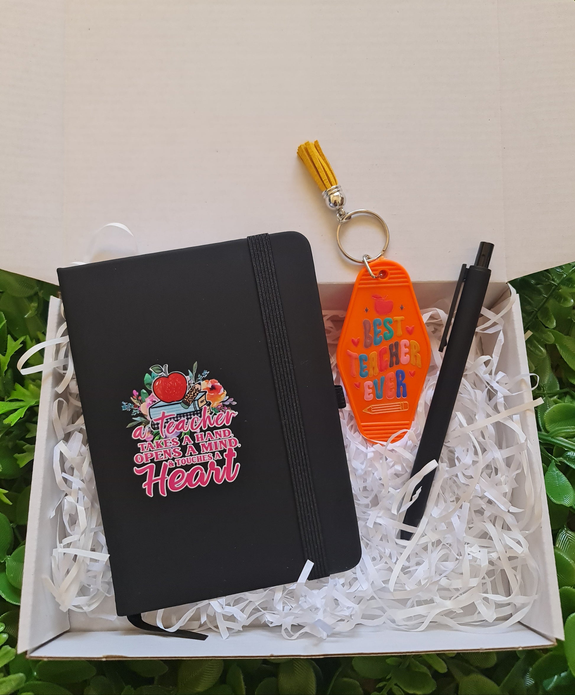 Black pouch with colorful text and graphics, orange keychain with colorful design, and black pen in a white box on grass.