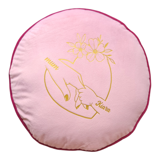 Round pink pillow with gold hand holding a flower design and text on a white background