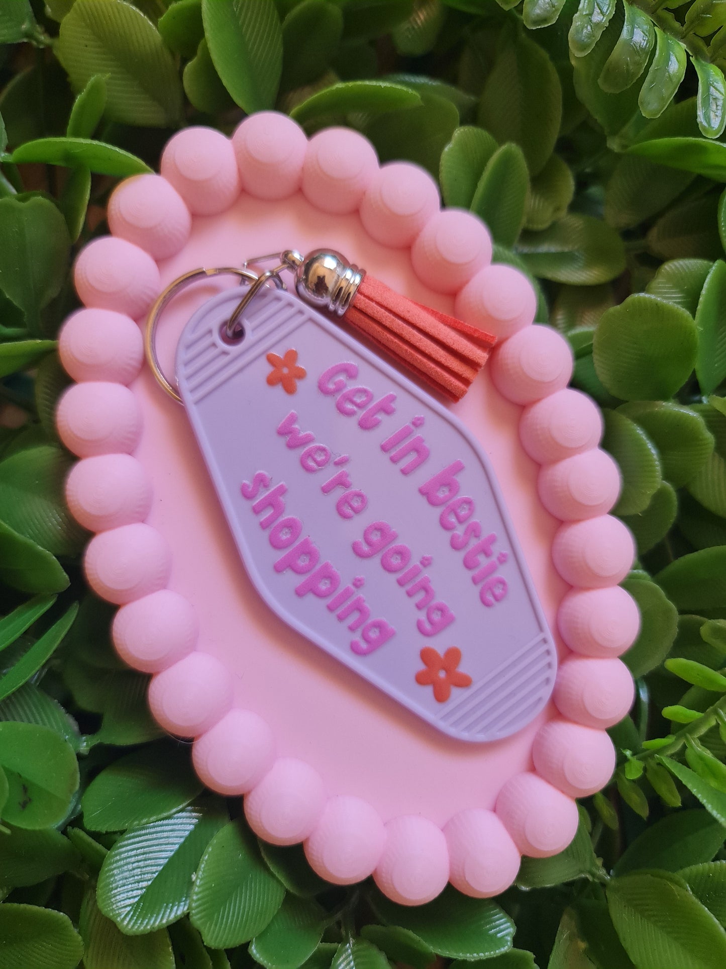 Pink keychain with text 'Get in bestie, we're going shopping' on a green leafy background