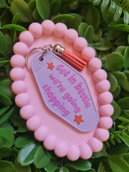 Pink keychain with text 'Get in bestie, we're going shopping' on a green leafy background