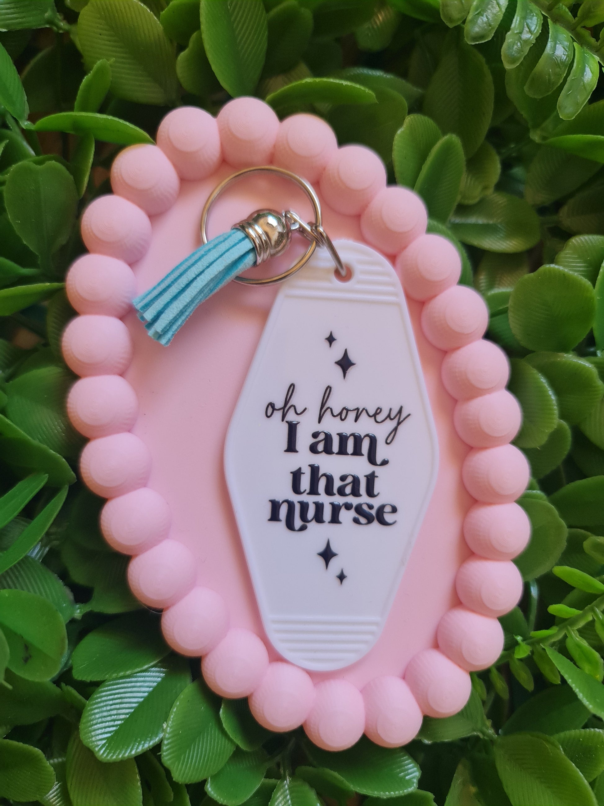 Pink keychain with a white tag and turquoise tassel on a green leafy background