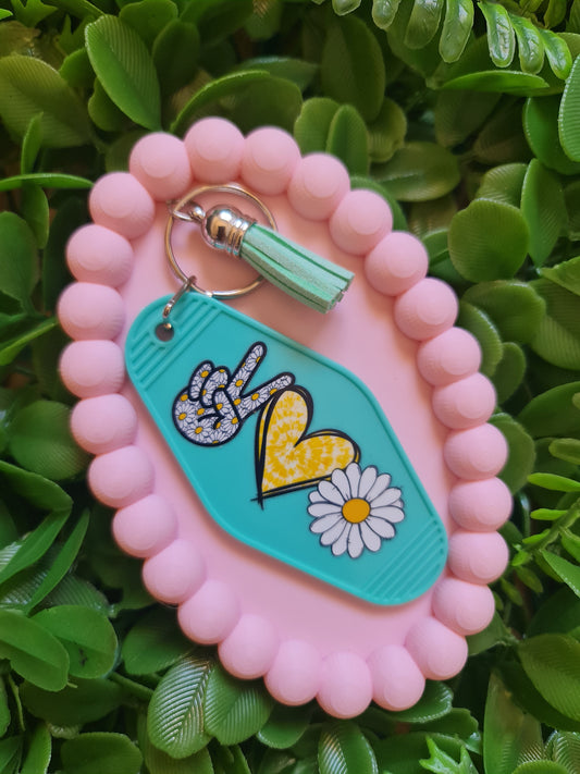 Pink keychain with a green tag featuring a peace sign and flower design, on a green leafy background.