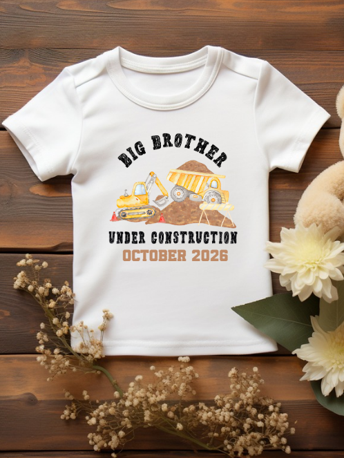 White t-shirt with 'Big Brother Under Construction October 2026' design, teddy bear, and flowers on a wooden background