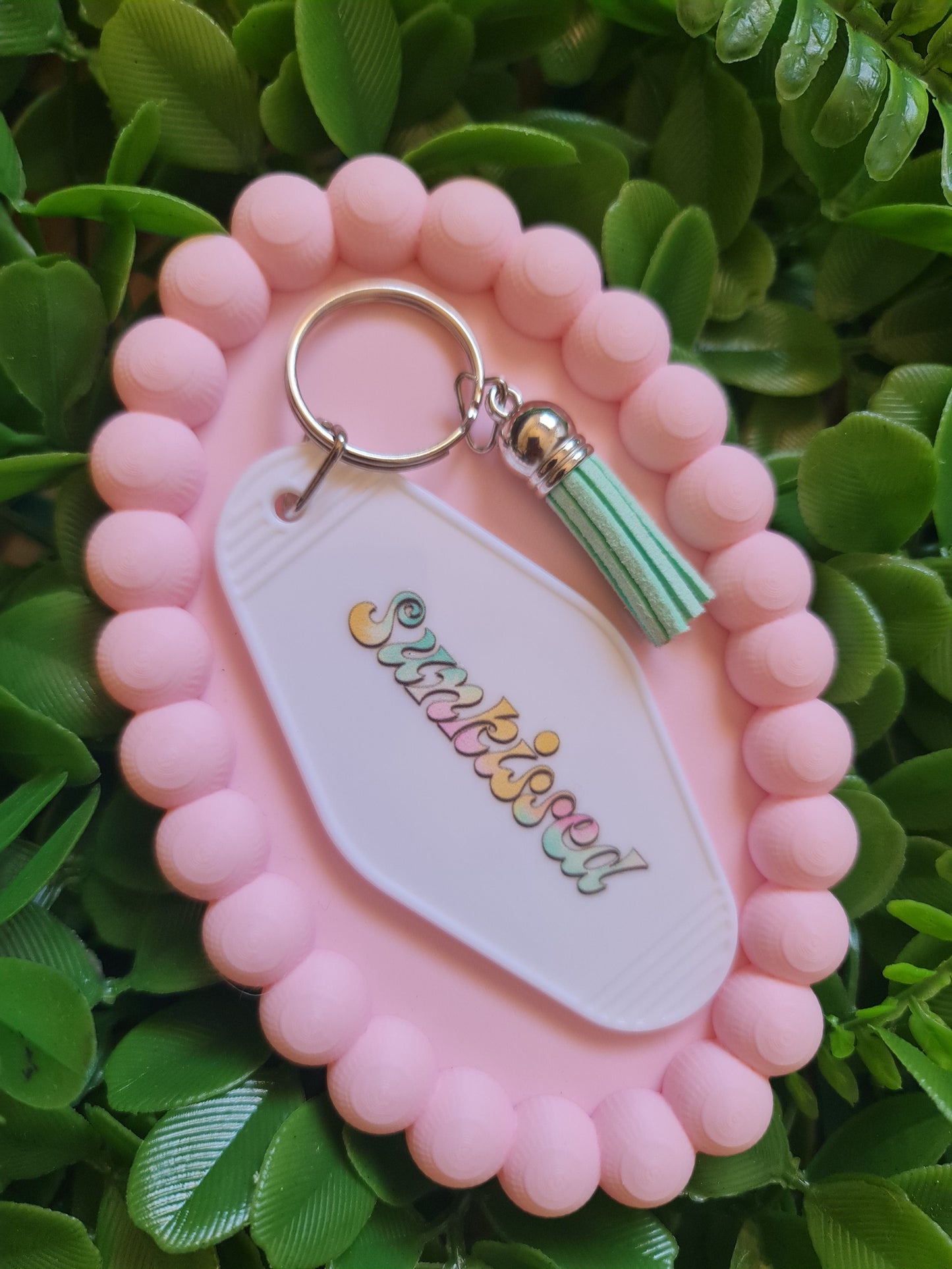 Pink keychain holder with a green tassel and 'Wishissimo' branding on a green leafy background