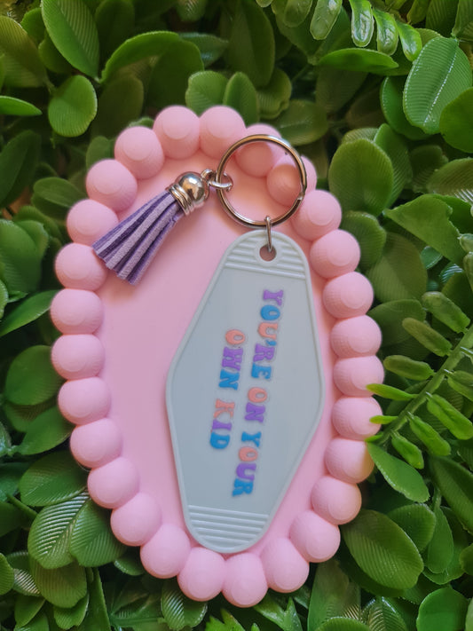 Pink keychain with a tag saying 'You're on your own kid' against a green leafy background