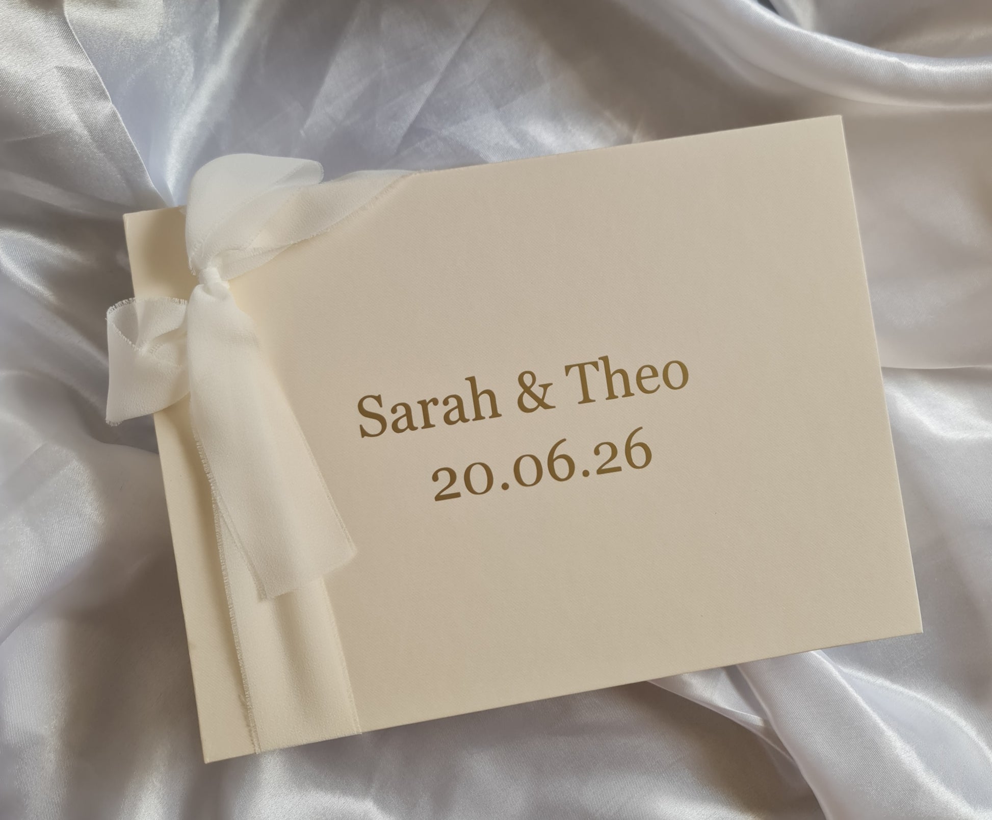 Beige card with 'Sarah & Theo' and a date on a white fabric background