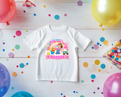 White t-shirt with colorful graphics and text on a wooden surface with balloons and confetti.