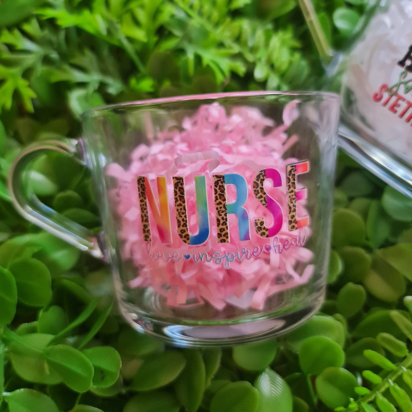 Clear mug with colorful text and pink decorative elements on a green leafy background