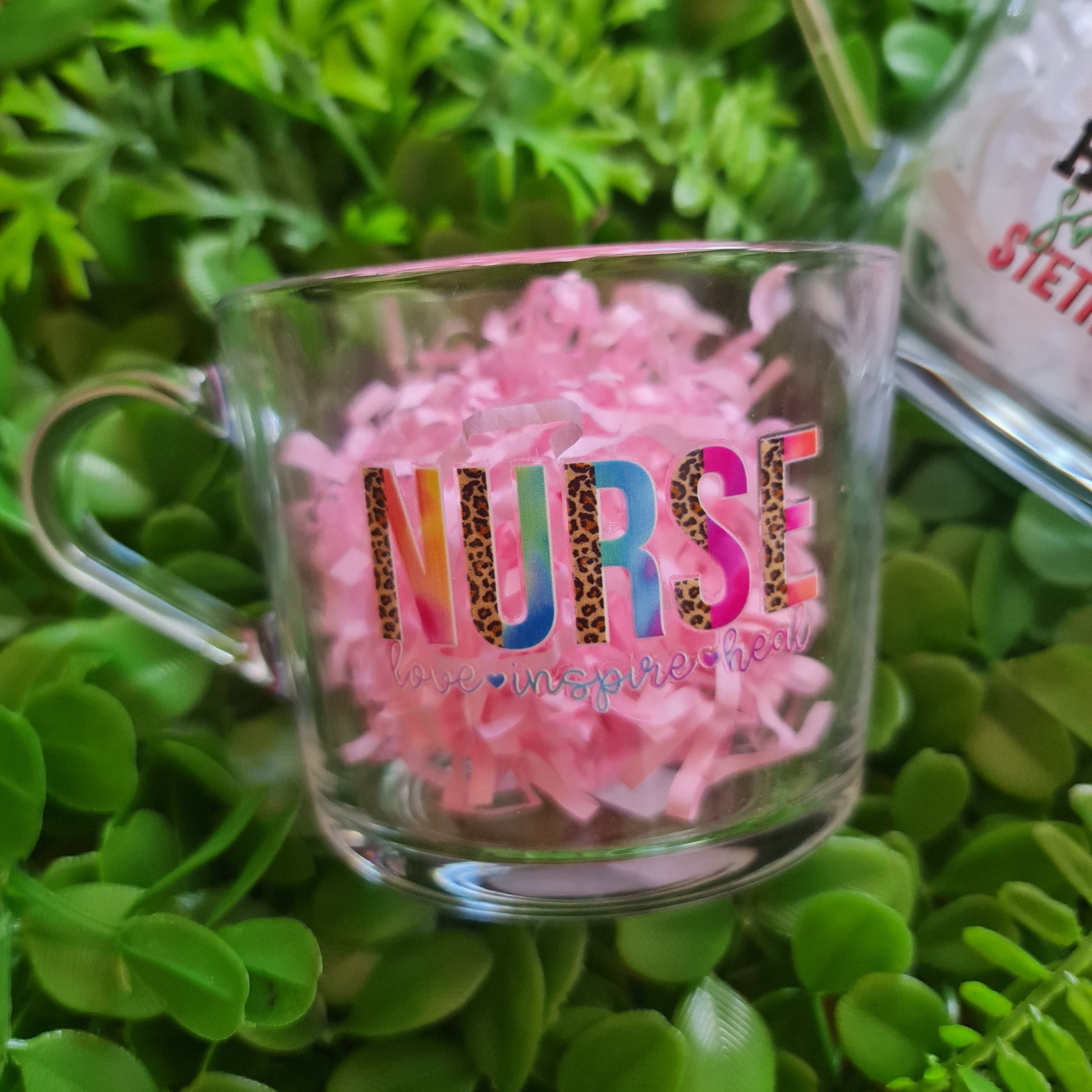 Clear mug with colorful text and pink decorative elements on a green leafy background
