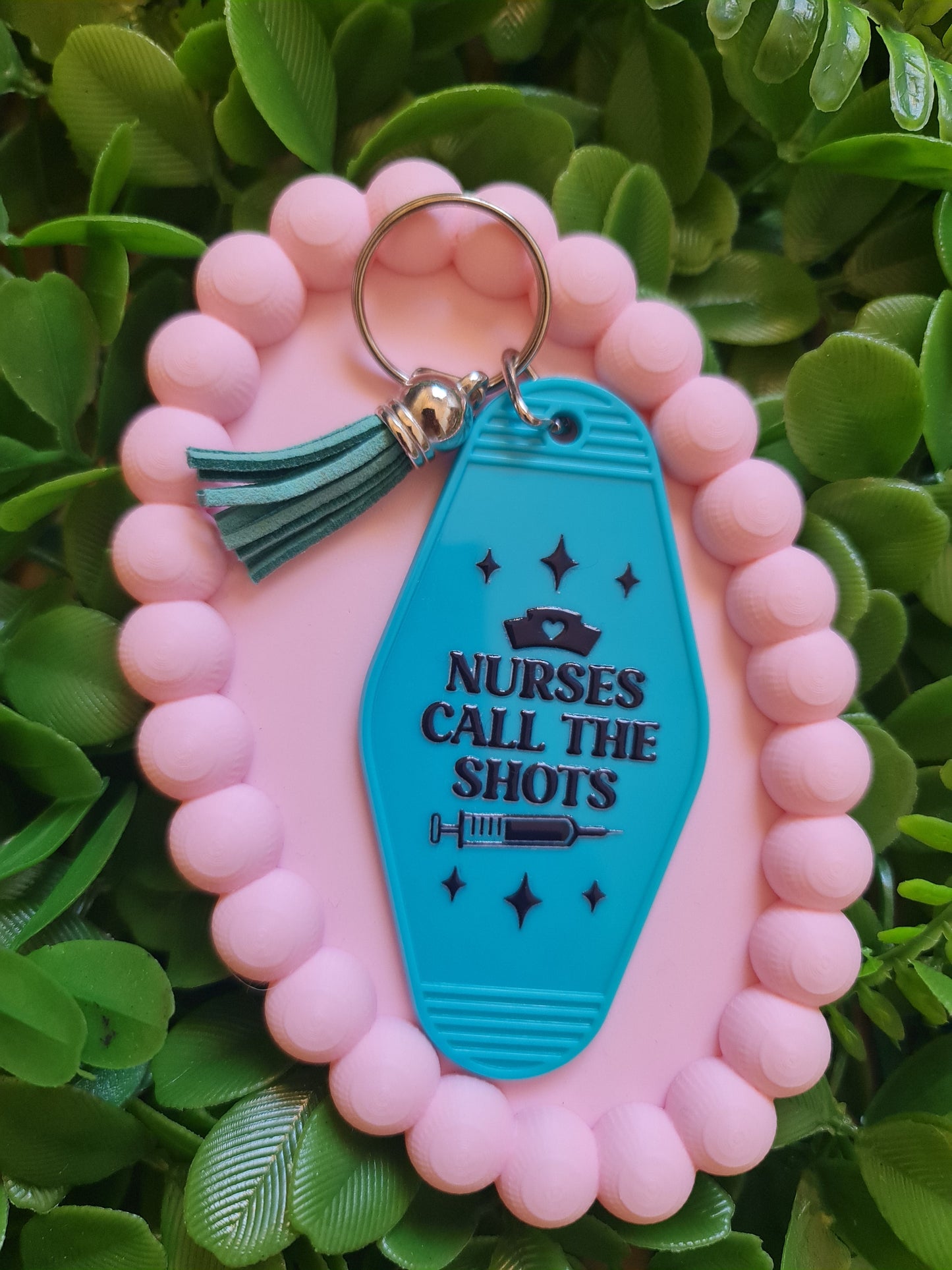 Pink keychain with a blue tag reading 'Nurses Call the Shots' on a green leafy background