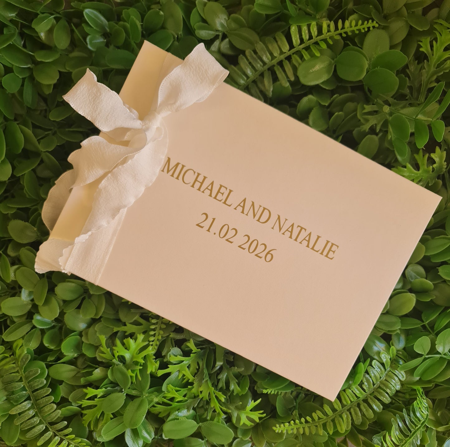 Beige wedding invitation with a white ribbon on green foliage