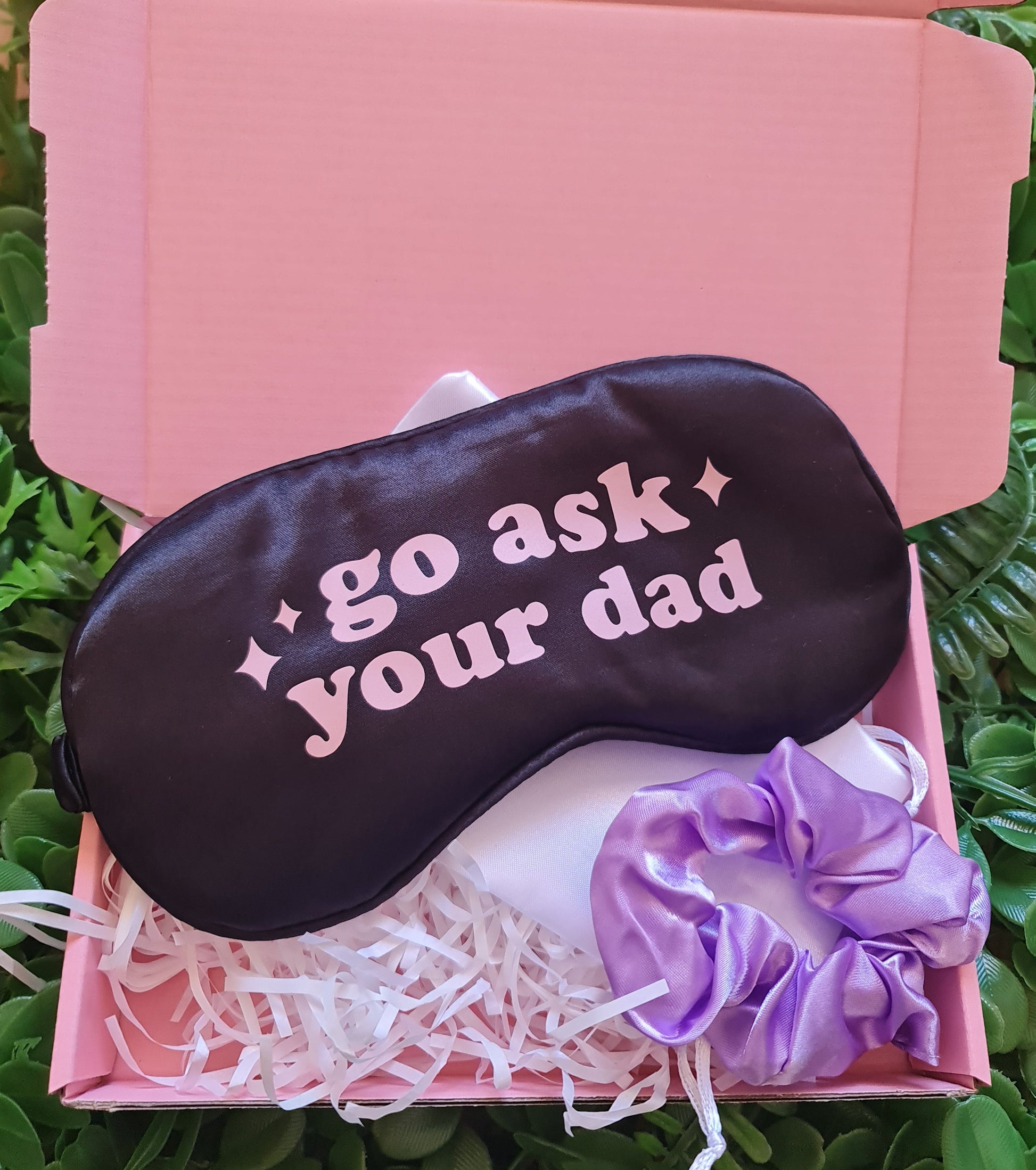 Sleep mask with text, white socks, and purple scrunchie in a pink box on green grass.