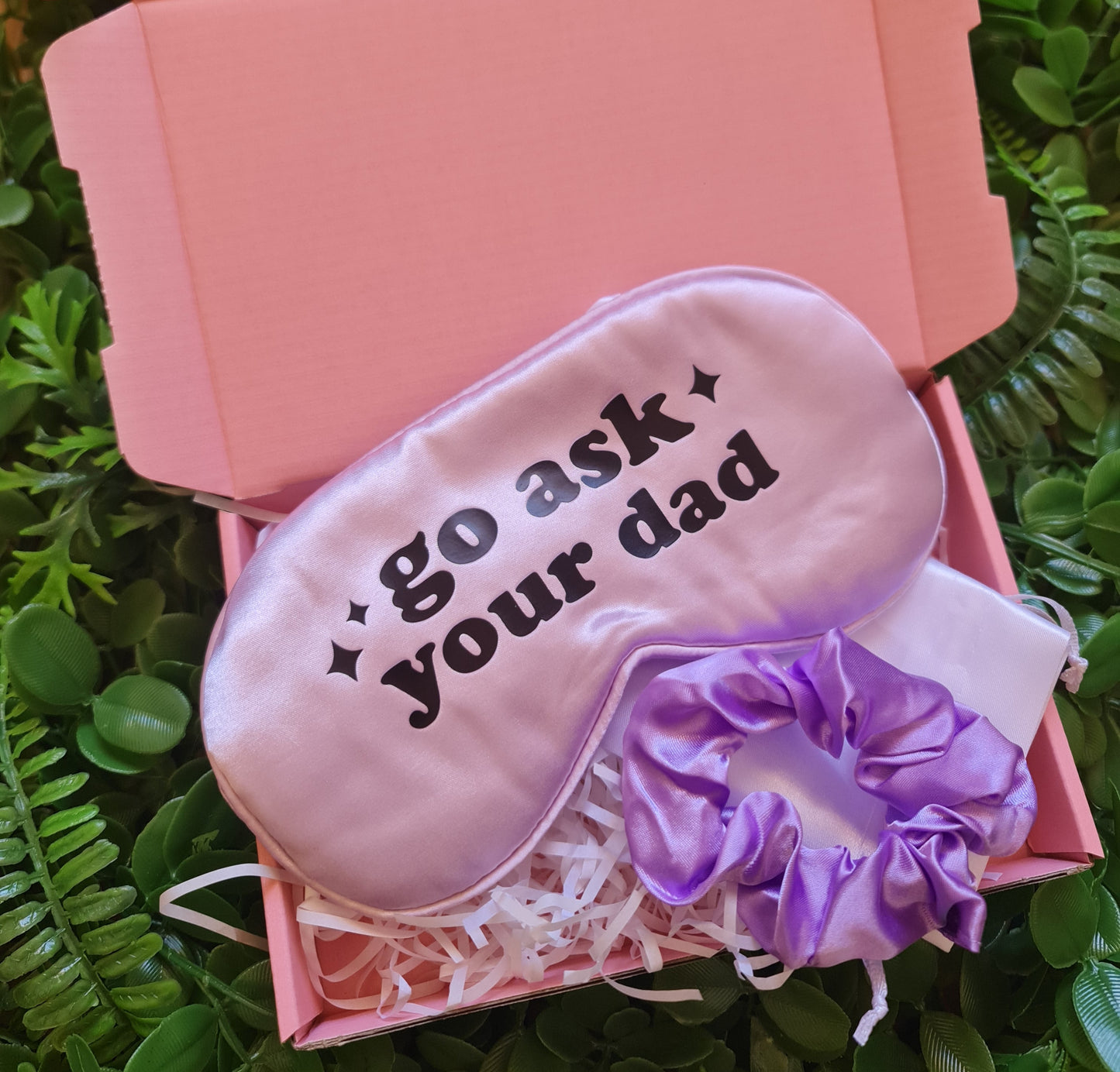 Pink sleep mask with text on a pink box with purple bow, placed on green grass.