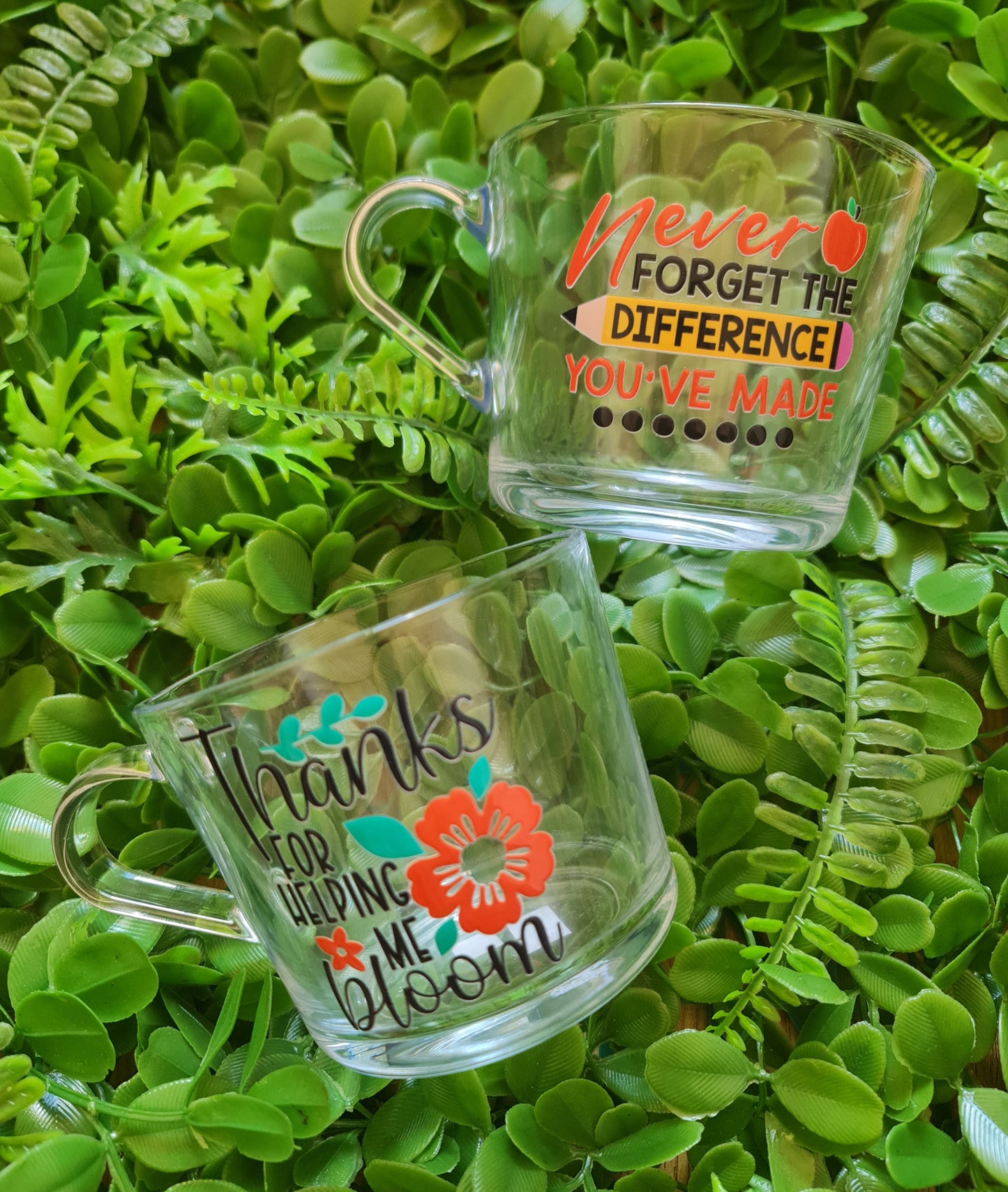 Two clear glass mugs with decorative text and designs on a green leafy background